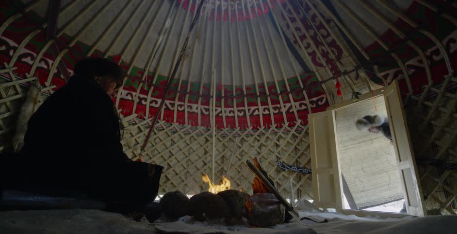 A mongolian man is entering a yurt where an older man is sitting by a fire