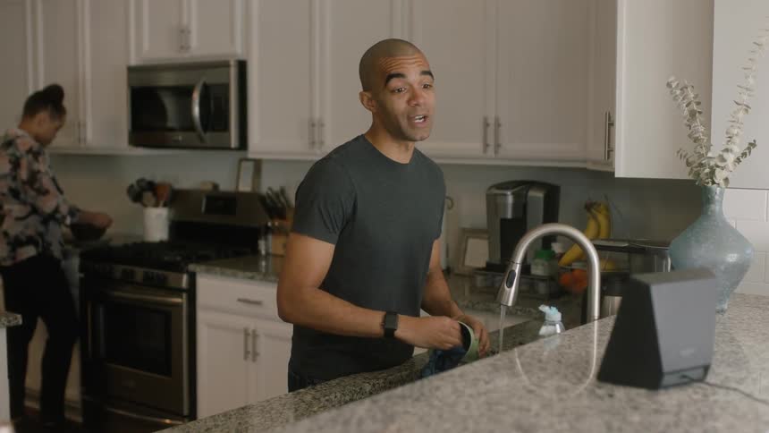 Man washes mug while talking to a woman cooking food in a kitchen