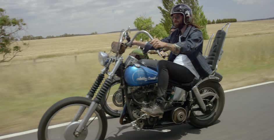 Men ride motorcycles past fields on a sunny day