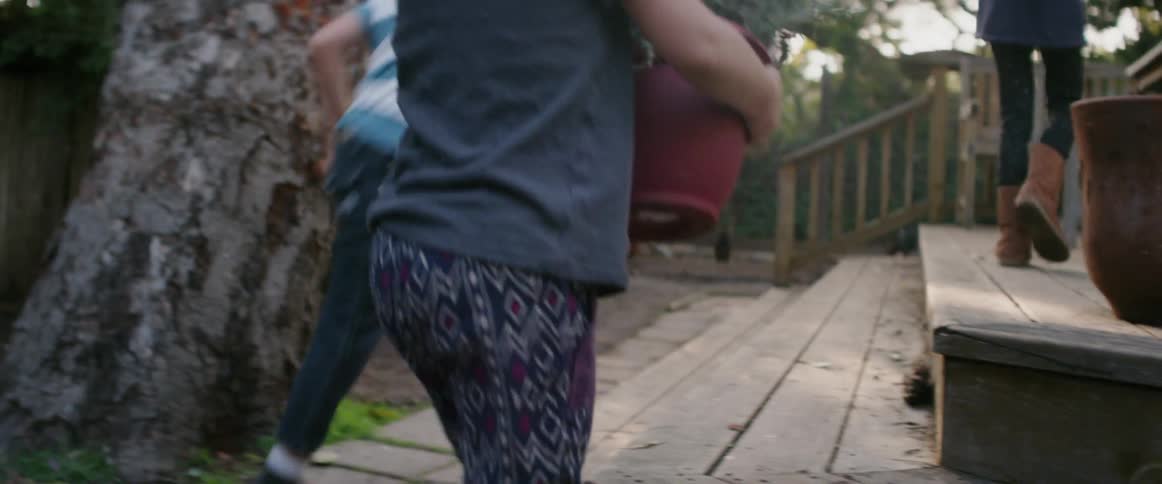 Three children are carrying potted plants onto a deck in their backyard in daylight