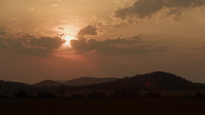 The sun is peeking through clouds as it sets over hills