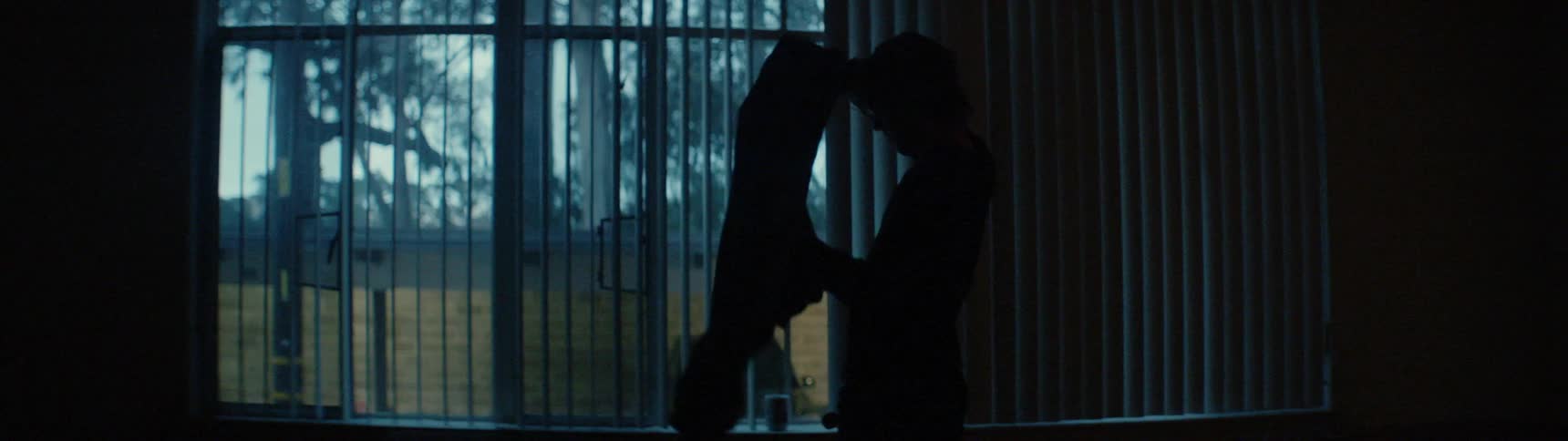 A man in a dark building picks up a jacket by a window and puts it on