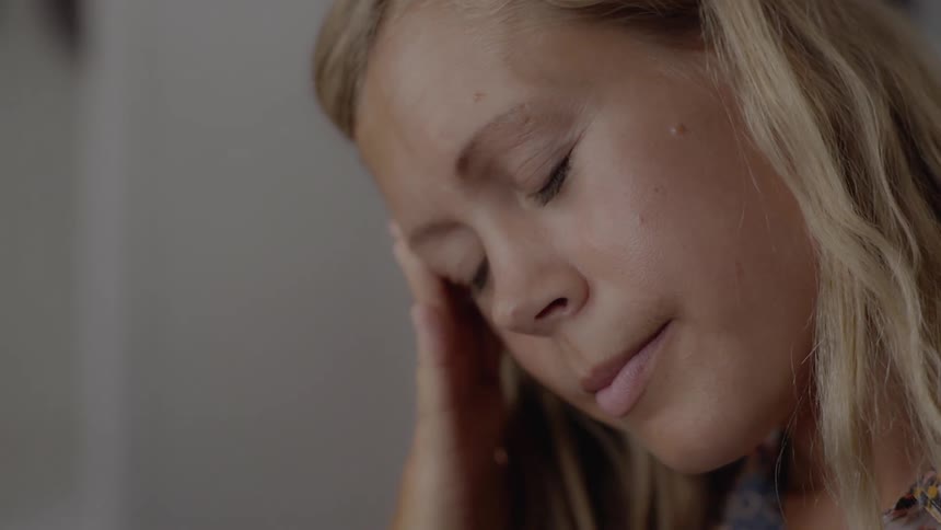A stressed blonde female closes her eyes while experiencing a painful headache indoors.