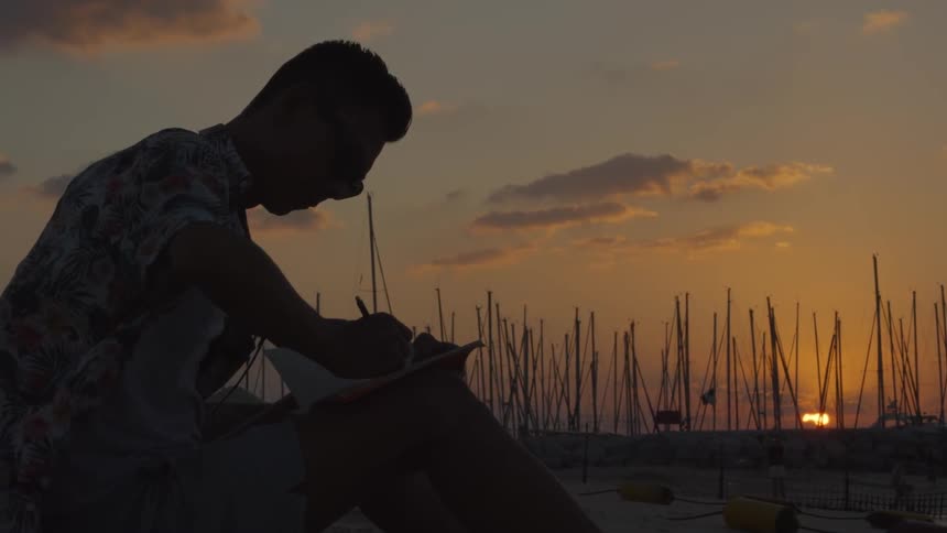A man writes in a notebook outside at sunset