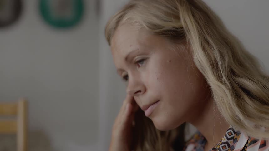 A focused blonde woman looks down thoughtfully while sitting alone inside her home