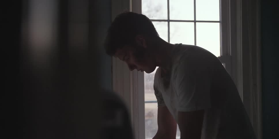 A boy with a white shirt bending down then standing up straight with a window in the background