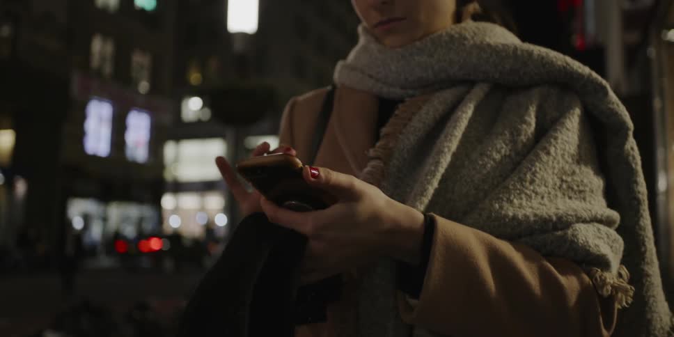 Woman standing on urban night sidewalk looks impatient and texts someone