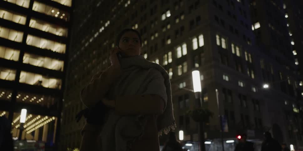A woman stands and uses her phone on a busy city sidewalk at night with large buildings behind her