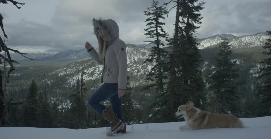 A woman and her corgi are walking on a snow-covered hillside in daylight