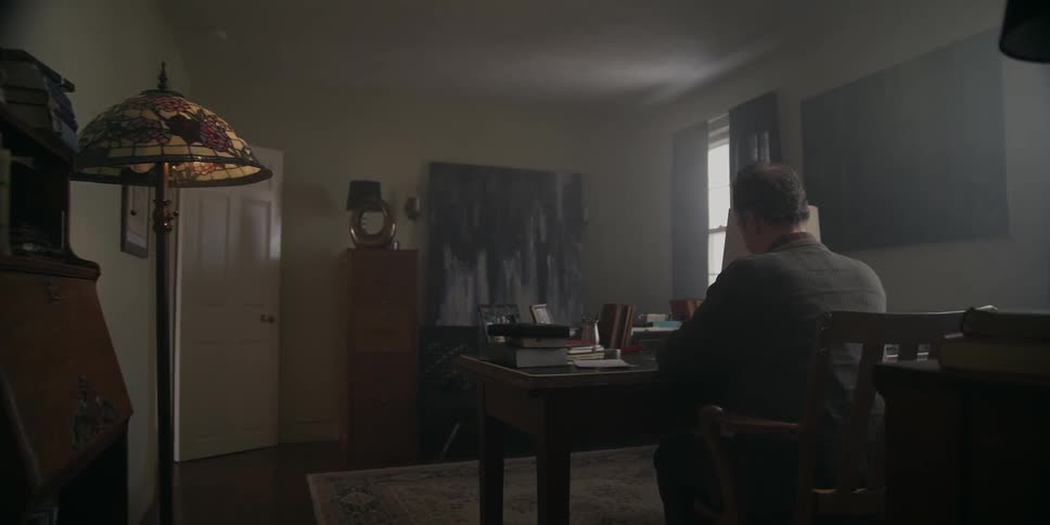 A man sitting at a desk in a dimly lit room looking at something he is holding in his hands