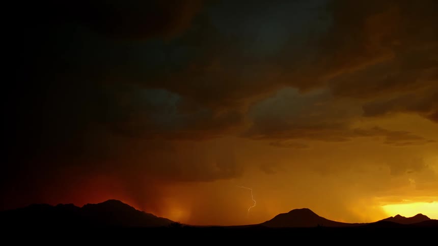 Lightning is flashing amid storm clouds as they move across a landscape at sunset