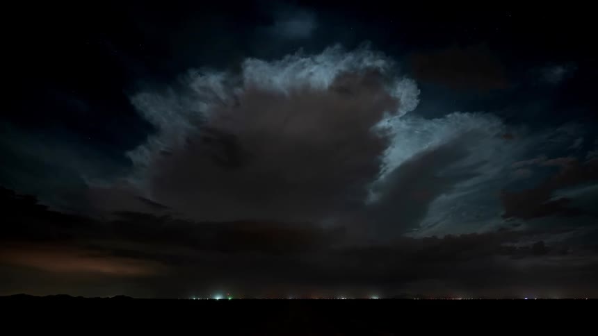 Lightning is flashing amid storm clouds as they move across a landscape at dusk