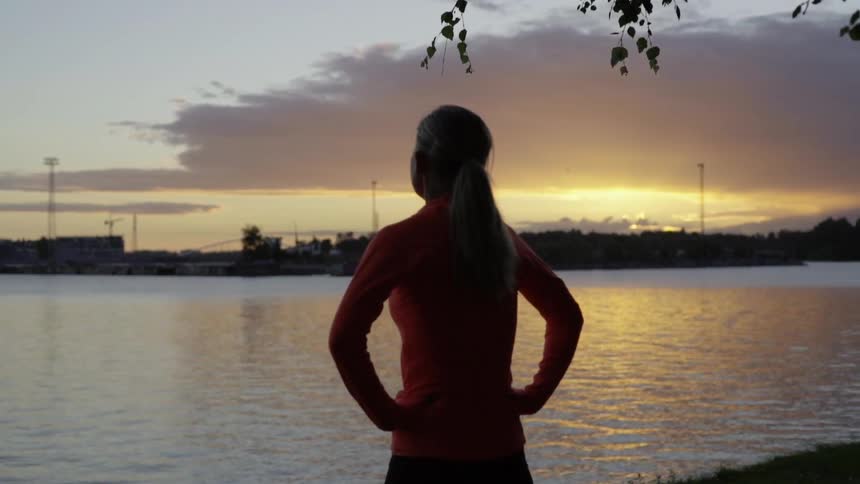 A woman in workout gear watches the sunset over the water