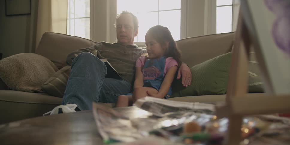 An older man sitting on a couch with a little girl as he reads to her
