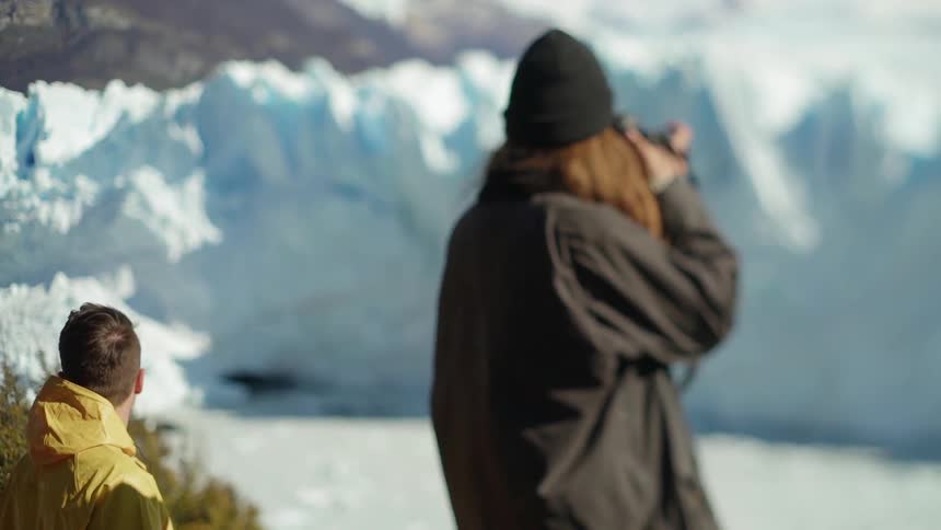 A man standing with a friend is taking a picture of a glacier on a sunny day