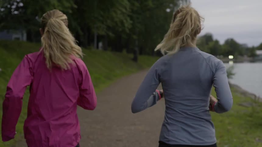Two blonde women in sportswear jog together along a scenic lakeside park trail.