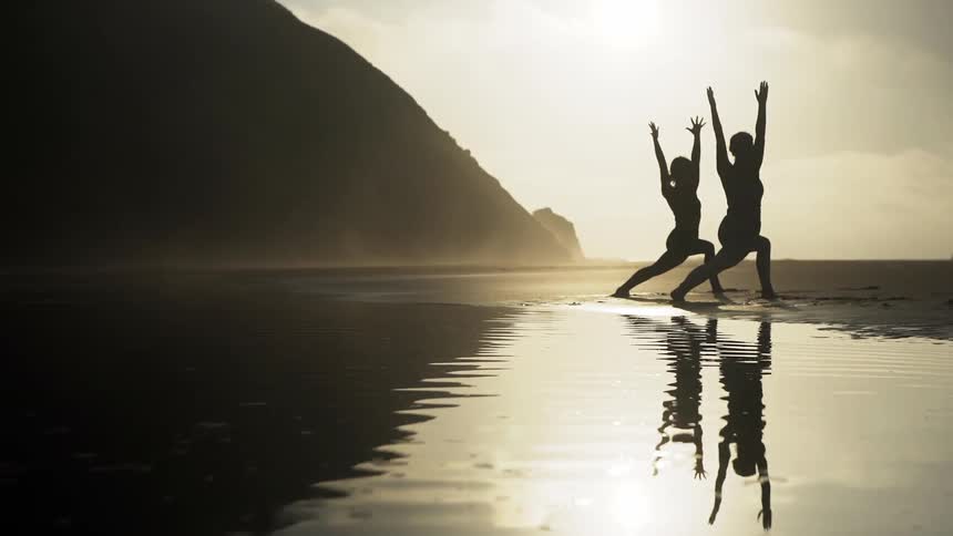 Two women do yoga on a beach at sunset