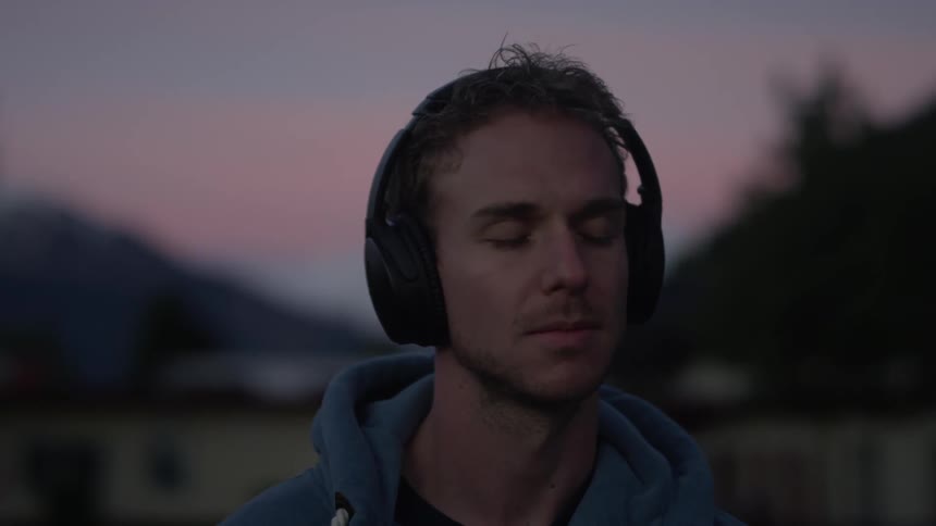 A man wearing headphones stands with his eyes closed with snow covered mountains and a pink sunset in the background