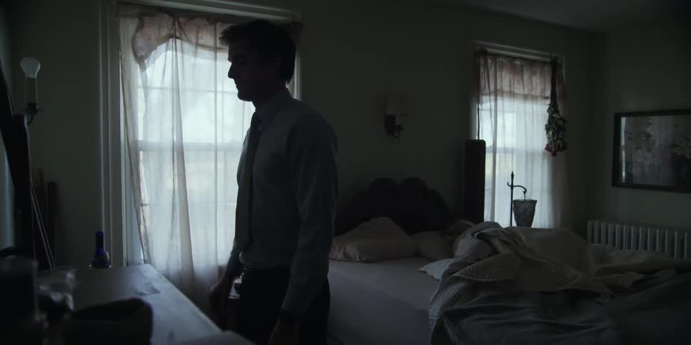 A man straightening his tie in a dresser mirror as a woman sleeps in the bed behind him