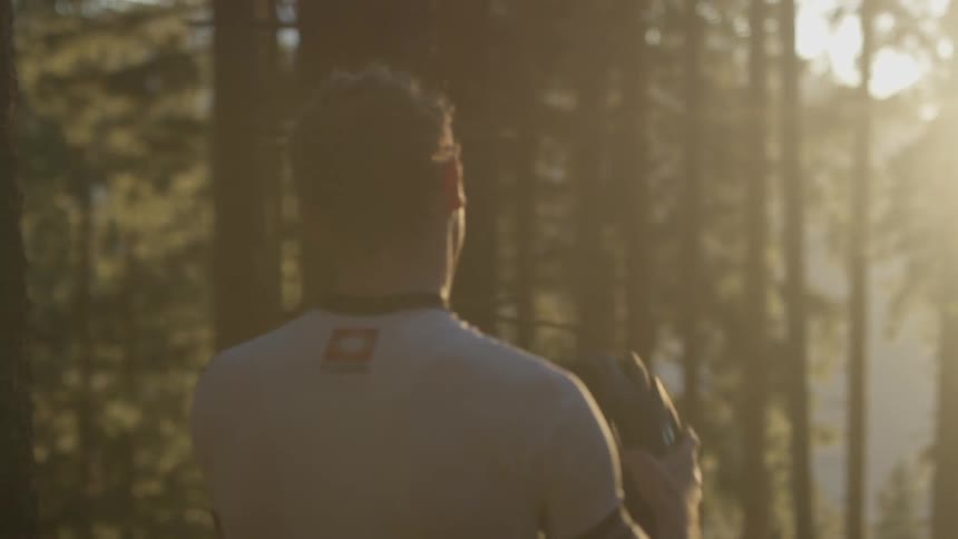 A man stands outside near some tall skinny trees on a sunny day and takes off his helmet