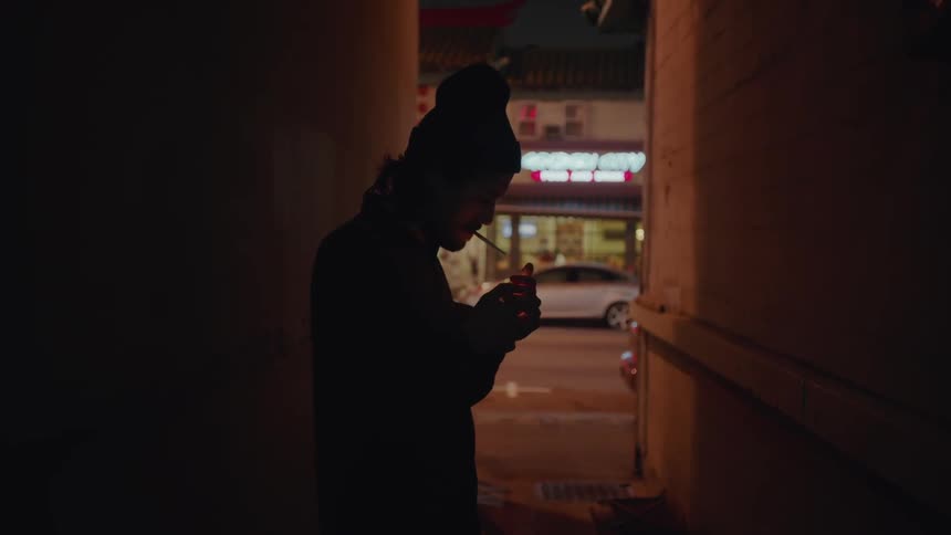 A man lights a cigarette as he walks in an alley at night