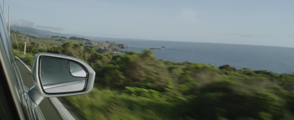 A vehicle is driving on a coastal road with a view of the ocean on a sunny day