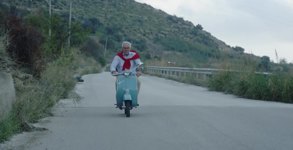 An older man is riding a vespa on a country road on a cloudy day