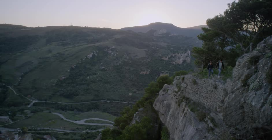 A couple are hiking along a cliff trail above a village at dawn