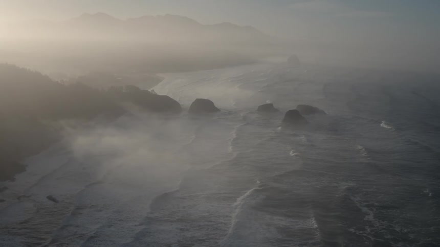 Waves are rolling toward a beach at the base of cliffs on a foggy morning