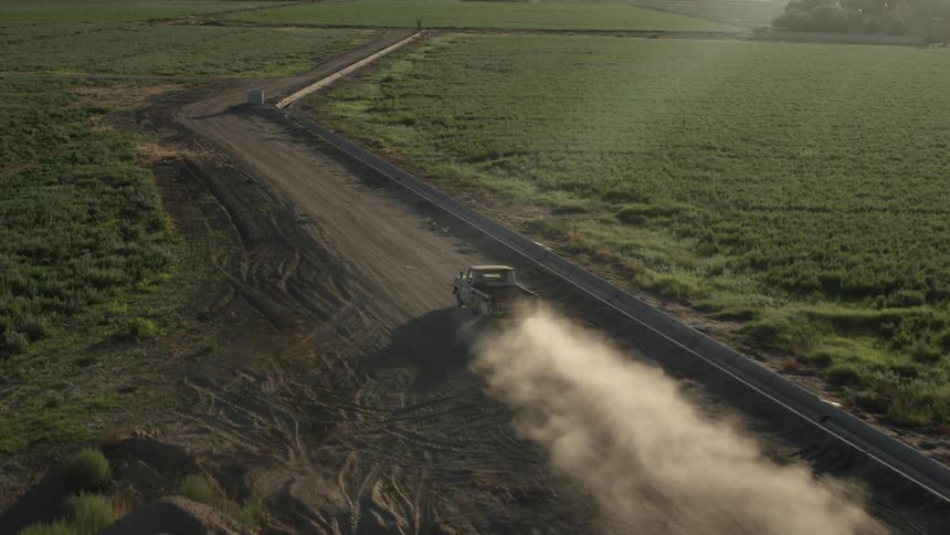 A pickup truck is kicking up clouds of dirt as it drives on a rural road in daylight