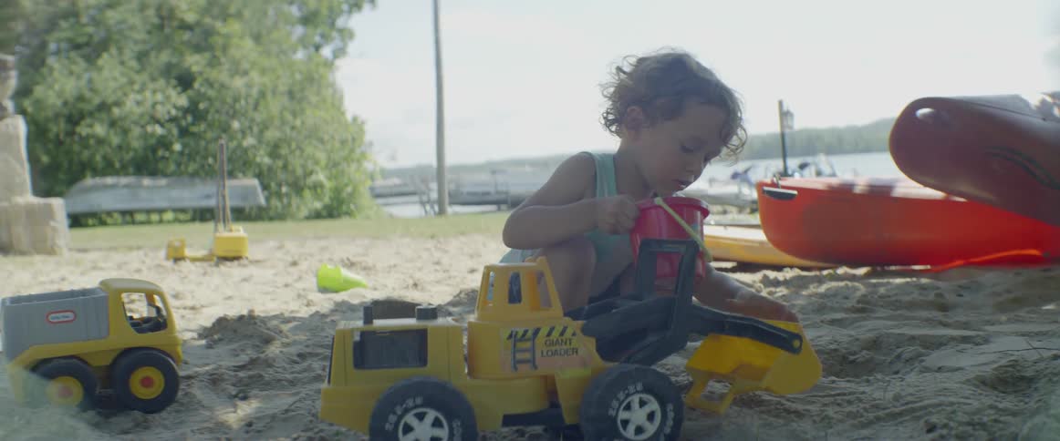 Boy in sandbox uses a bucket to fill toy backhoe with sand