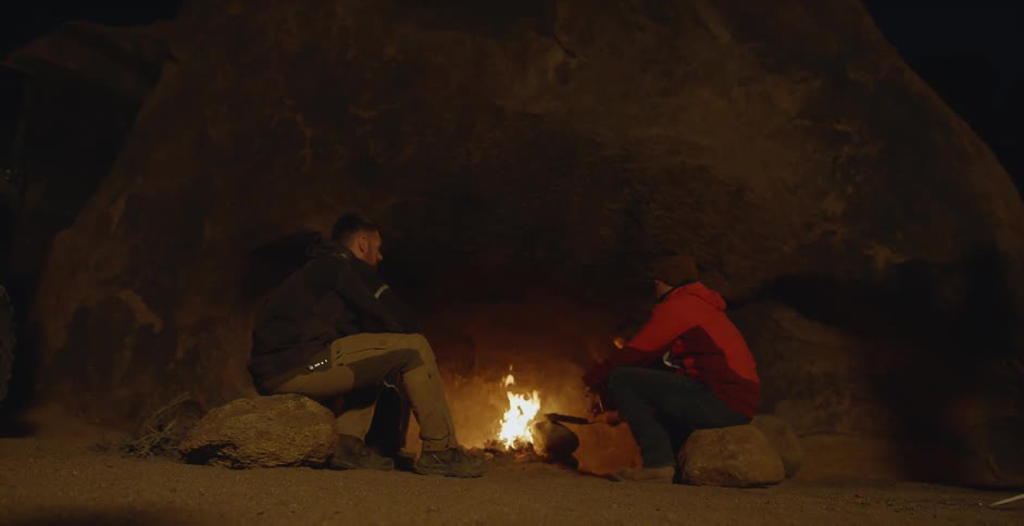 A couple are sitting and cooking at a campfire outside at night