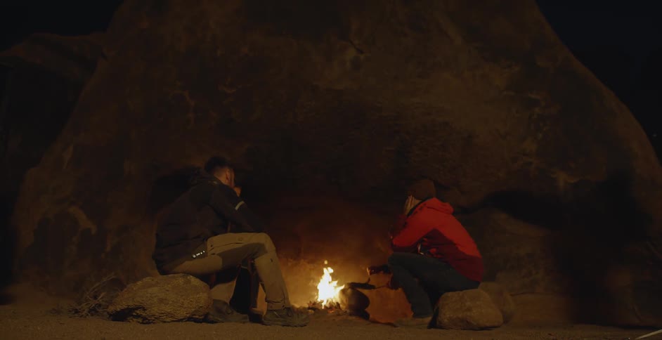 Two men sit by a campfire at night
