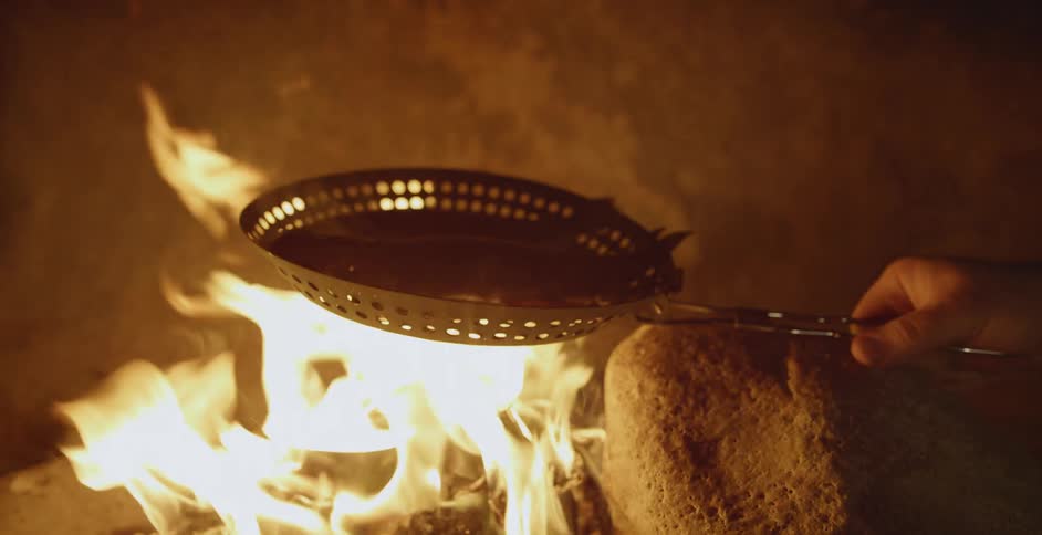 A pan with fish in it is held over an open campfire