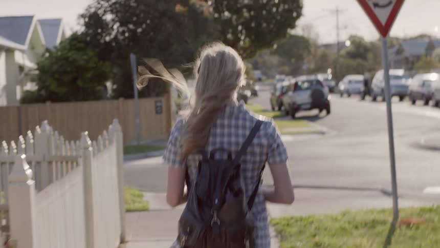 A young, blonde woman wearing a backpack and walking down a neighborhood street as the wind blows her hair