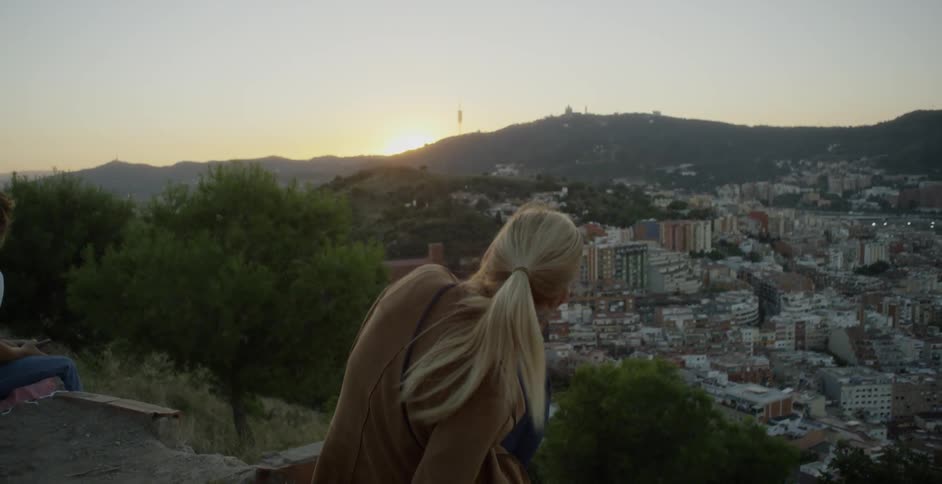 A woman in a brown jacket is sitting on the edge of a hillside overlooking the city