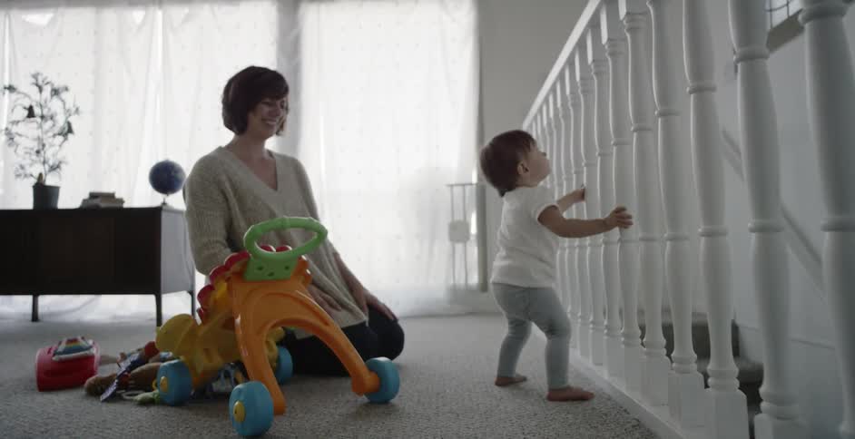 A baby is standing up against a banister while its mommy is watching
