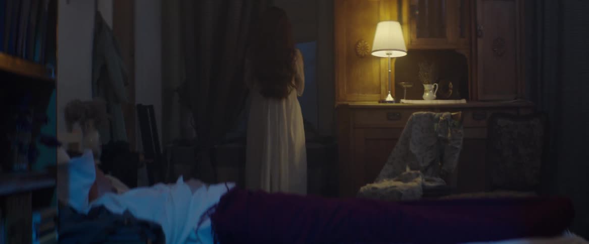 Woman standing and looking out a dark window with a lamp barely lighting the bedroom