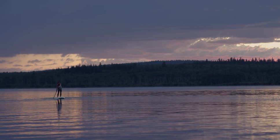 Woman paddling on paddle board at dusk while lightning strikes in cloudy sky