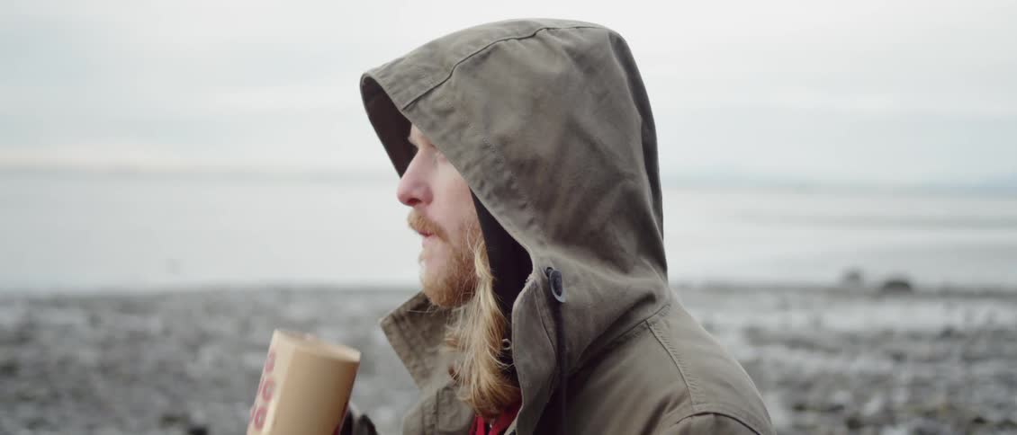 Bearded man drinking from a mug near the sea on a chilly day wearing a hoodie