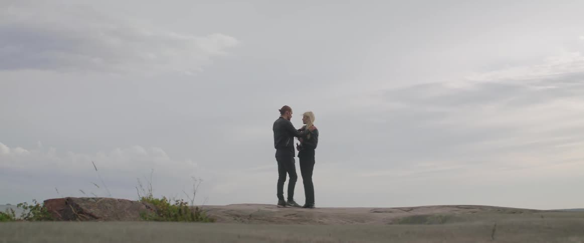 Two lovers embrace gently while standing together under a cloudy grey sky outdoors.