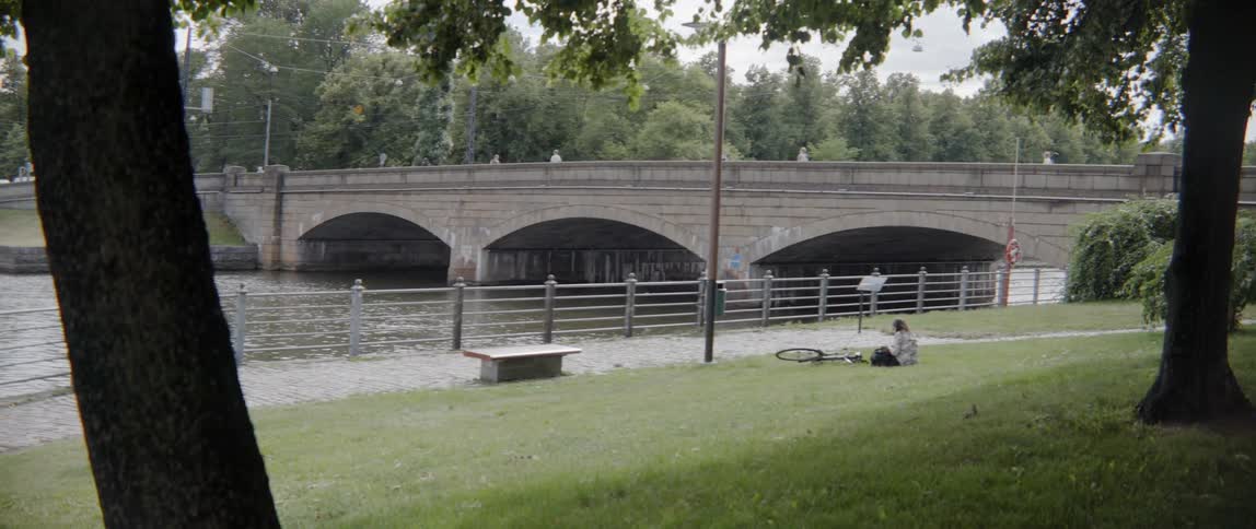 Distant people walk across a concrete bridge over a river next to park.
