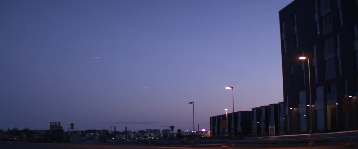 Tall lampposts glow brightly along an empty city street during a purple sunset.