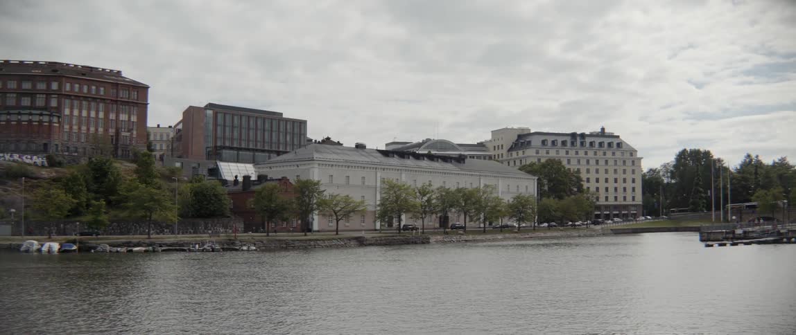 Green trees line a peaceful urban waterfront under an overcast summer sky outdoors.