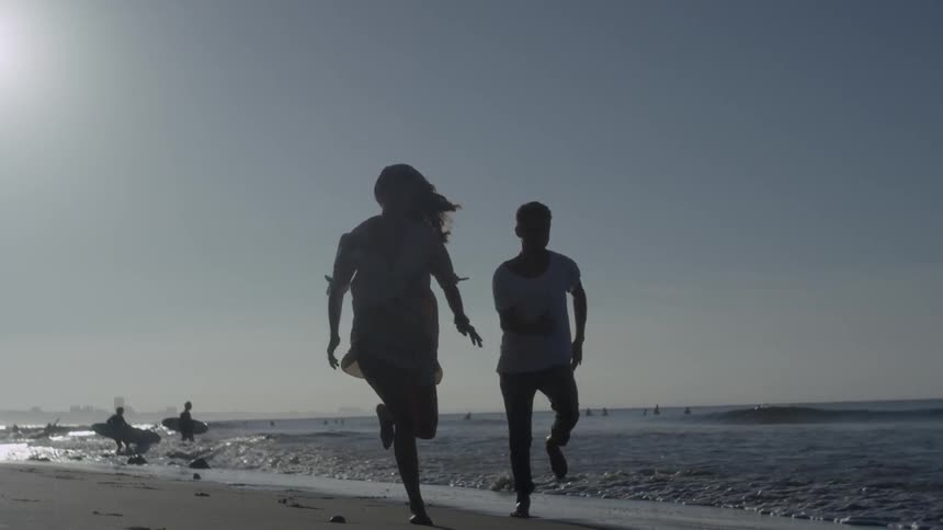 Man and woman racing on a beach as others carry their surfboards into the waters