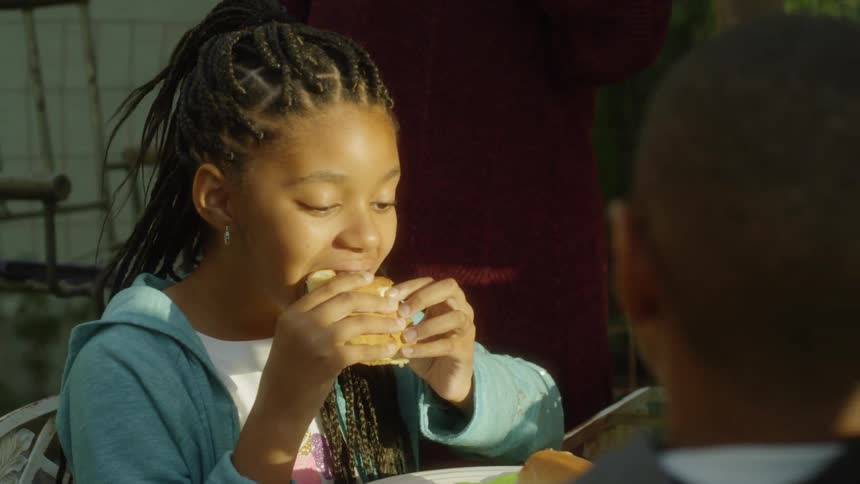 A girl sitting at an outdoor table with family takes a bite of her sandwich