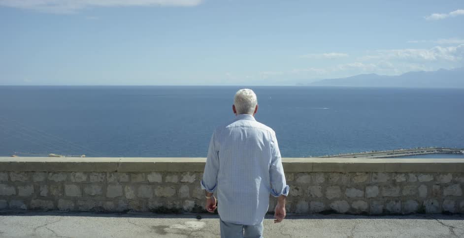 An older man is walking toward a wall with a view of the ocean on a sunny day
