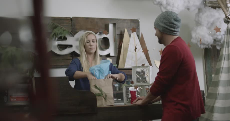 A woman in a shop wraps and bags an item and then hands it to a man