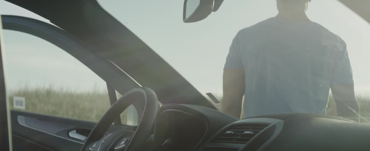 A man is leaning against the hood of a vehicle parked near a field on a sunny day