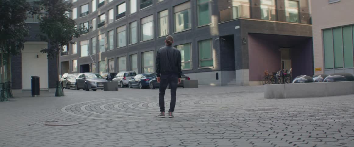 A young man looks at contemporary corporate buildings and parked cars in plaza.